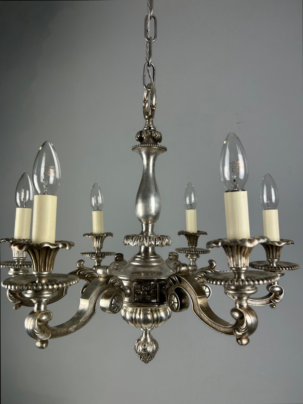 An antique silver-plated six-light chandelier featuring curved scrolling arms, gadrooned drip pans, cream candle sleeves, and a central baluster stem. View II