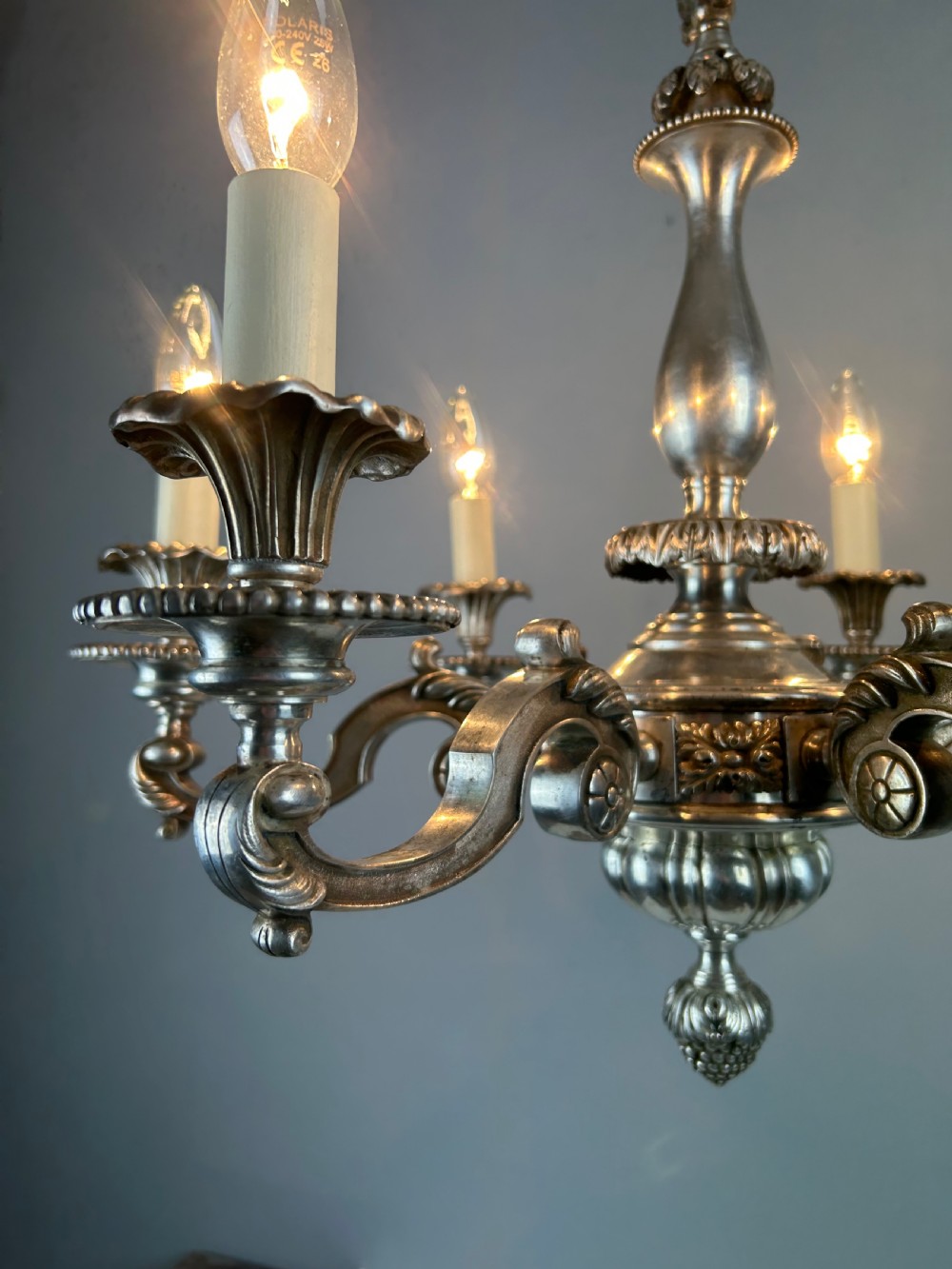 An antique silver-plated six-light chandelier featuring curved scrolling arms, gadrooned drip pans, cream candle sleeves, and a central baluster stem. View III
