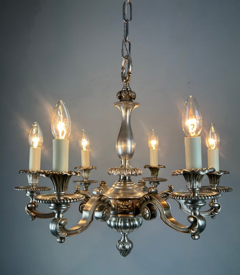An antique silver-plated six-light chandelier featuring curved scrolling arms, gadrooned drip pans, cream candle sleeves, and a central baluster stem. View V