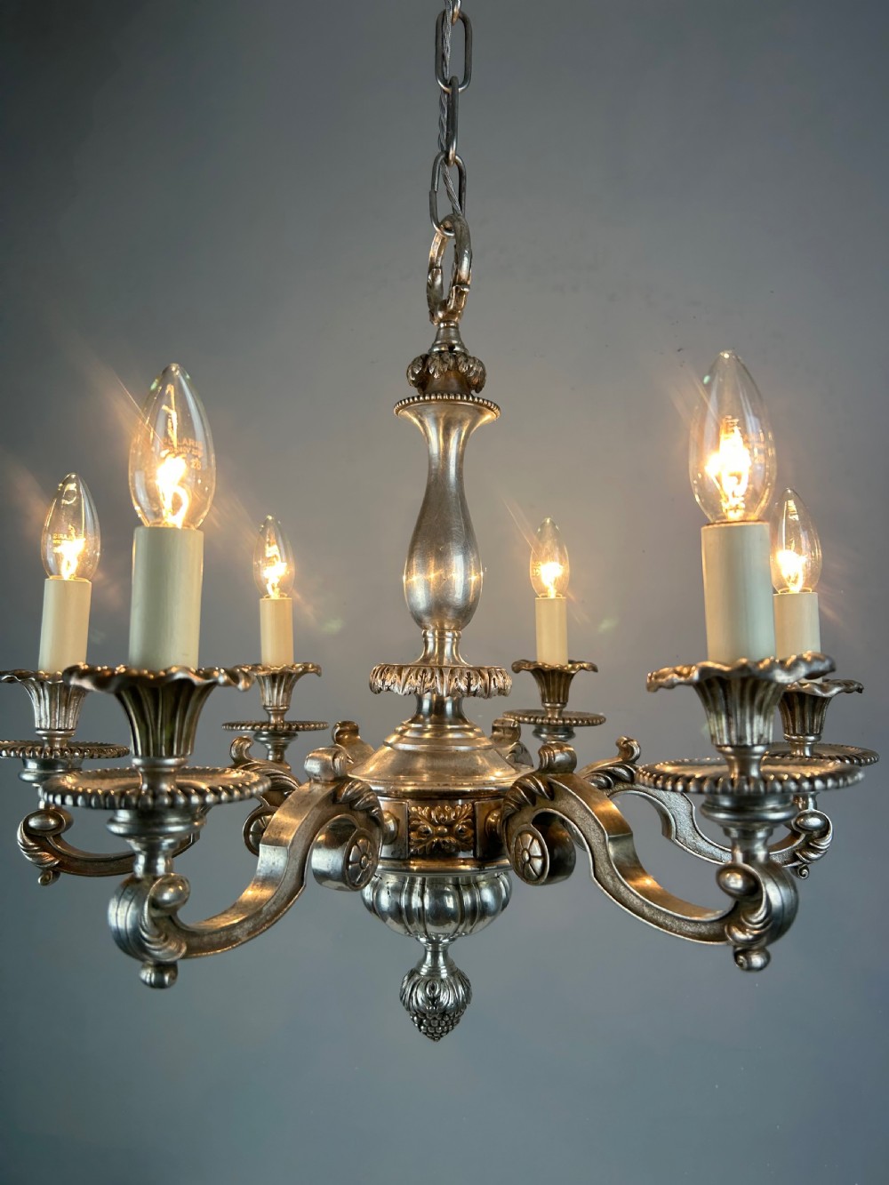 An antique silver-plated six-light chandelier featuring curved scrolling arms, gadrooned drip pans, cream candle sleeves, and a central baluster stem. View VII