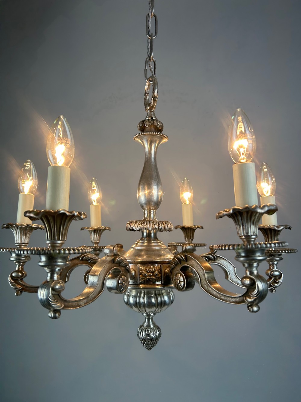 An antique silver-plated six-light chandelier featuring curved scrolling arms, gadrooned drip pans, cream candle sleeves, and a central baluster stem. View VIII