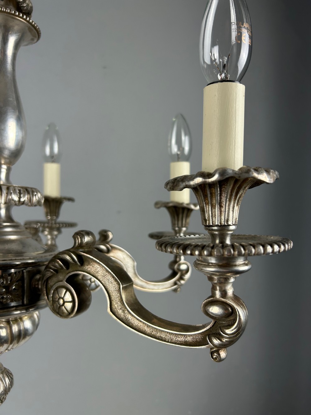An antique silver-plated six-light chandelier featuring curved scrolling arms, gadrooned drip pans, cream candle sleeves, and a central baluster stem. View IX