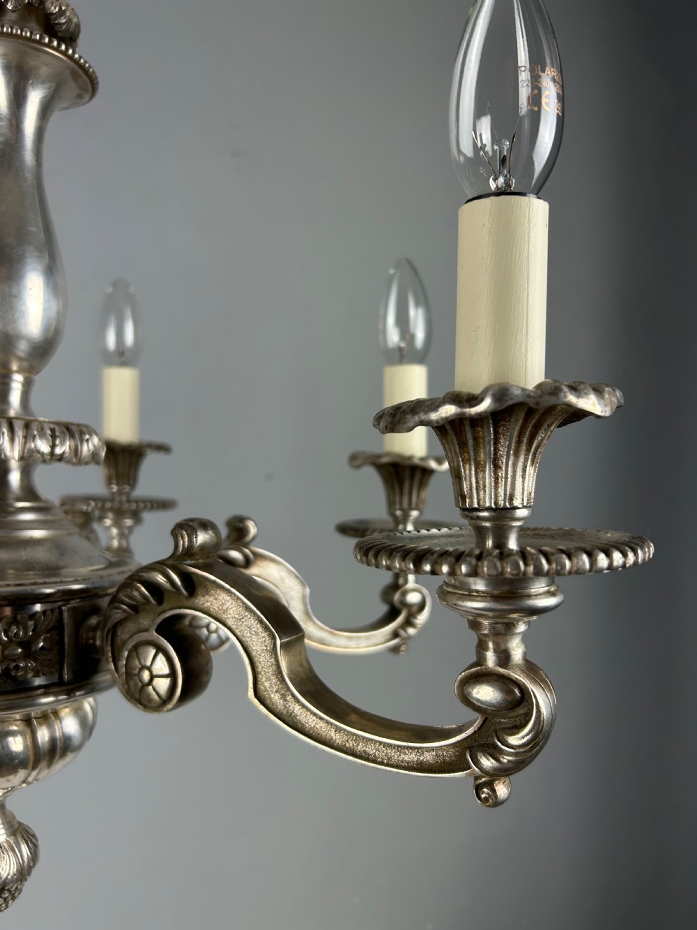 An antique silver-plated six-light chandelier featuring curved scrolling arms, gadrooned drip pans, cream candle sleeves, and a central baluster stem. View X