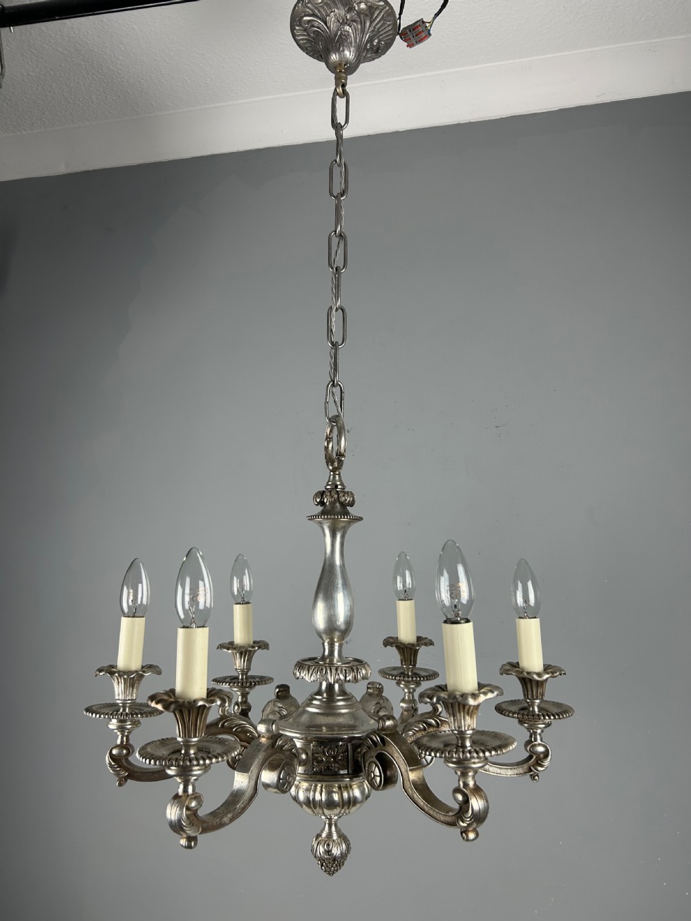 An antique silver-plated six-light chandelier featuring curved scrolling arms, gadrooned drip pans, cream candle sleeves, and a central baluster stem. View XI