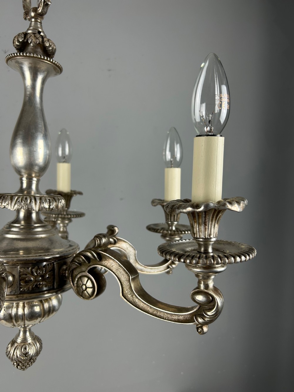 An antique silver-plated six-light chandelier featuring curved scrolling arms, gadrooned drip pans, cream candle sleeves, and a central baluster stem. View XII