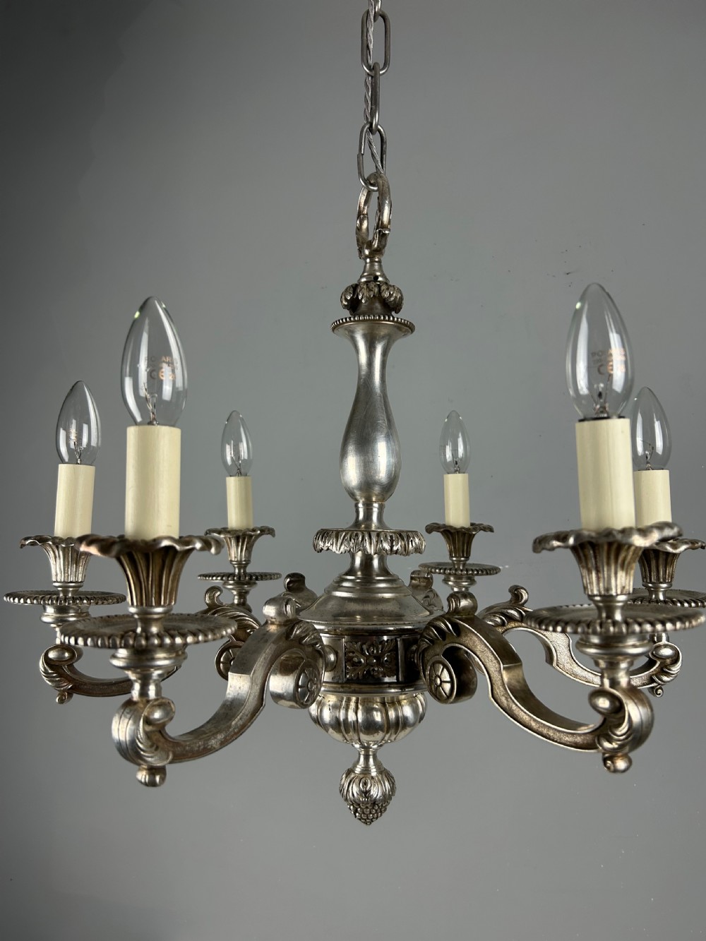 An antique silver-plated six-light chandelier featuring curved scrolling arms, gadrooned drip pans, cream candle sleeves, and a central baluster stem. View XIII
