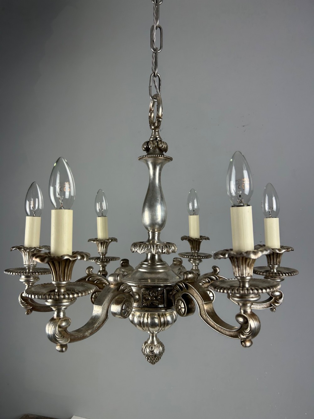 An antique silver-plated six-light chandelier featuring curved scrolling arms, gadrooned drip pans, cream candle sleeves, and a central baluster stem. View XVI