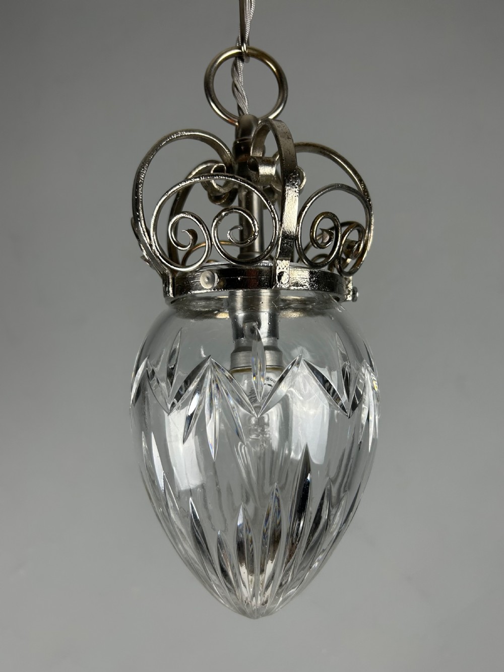 A clear cut glass acorn-shaped shade topped with a scrollwork chrome crown gallery and suspended wire.
