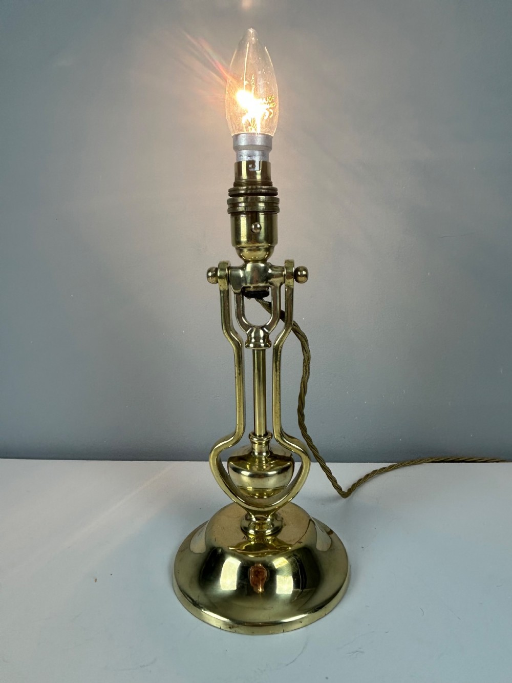 An antique brass gimbal table lamp featuring a circular weighted base, pivoted swinging frame, and exposed bayonet bulb socket. View II