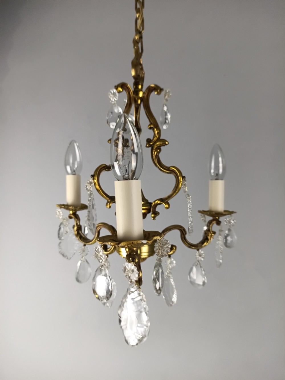 A French gilt brass three-arm chandelier featuring curved scrollwork arms, candle-style lamp holders, and dangling faceted crystal droplets. View X