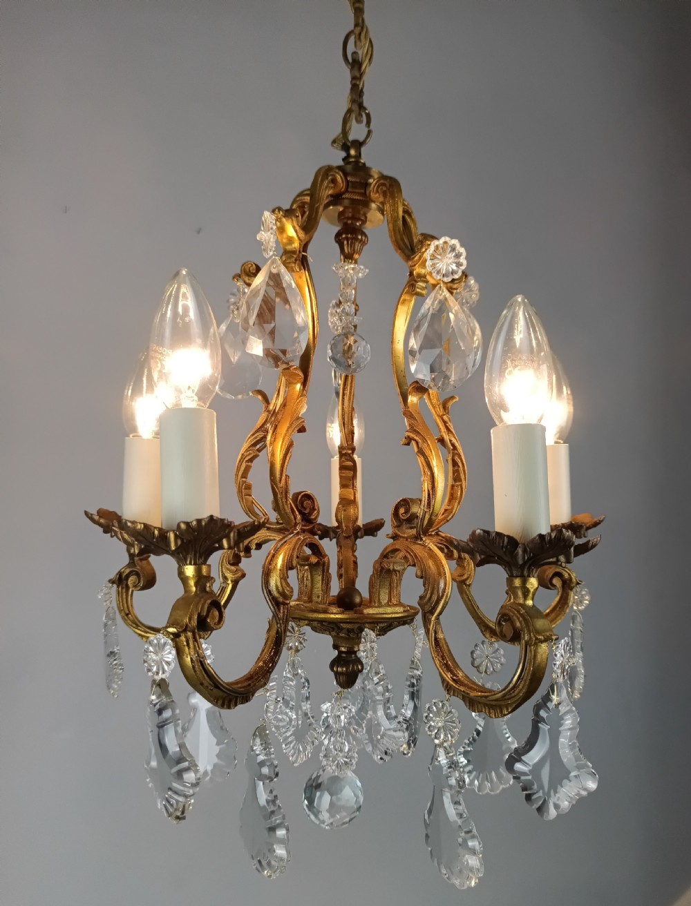 A five-arm French Rococo style chandelier featuring a gilded scrollwork frame adorned with clear faceted crystal drops and white candle sleeves. View II
