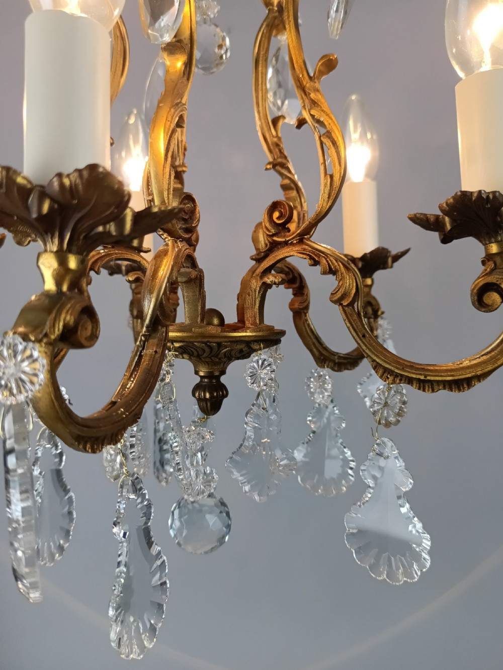 A five-arm French Rococo style chandelier featuring a gilded scrollwork frame adorned with clear faceted crystal drops and white candle sleeves. View IV
