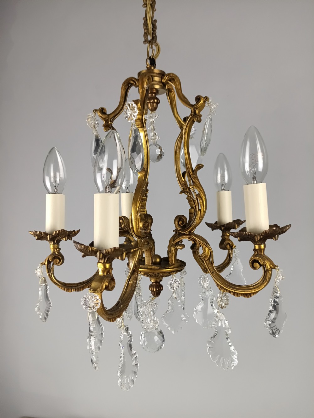 A five-arm French Rococo style chandelier featuring a gilded scrollwork frame adorned with clear faceted crystal drops and white candle sleeves. View VIII