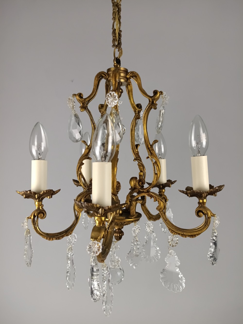 A five-arm French Rococo style chandelier featuring a gilded scrollwork frame adorned with clear faceted crystal drops and white candle sleeves. View IX
