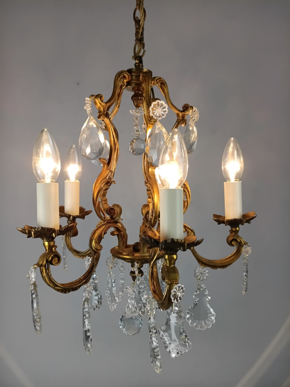 A five-arm French Rococo style chandelier featuring a gilded scrollwork frame adorned with clear faceted crystal drops and white candle sleeves. View X