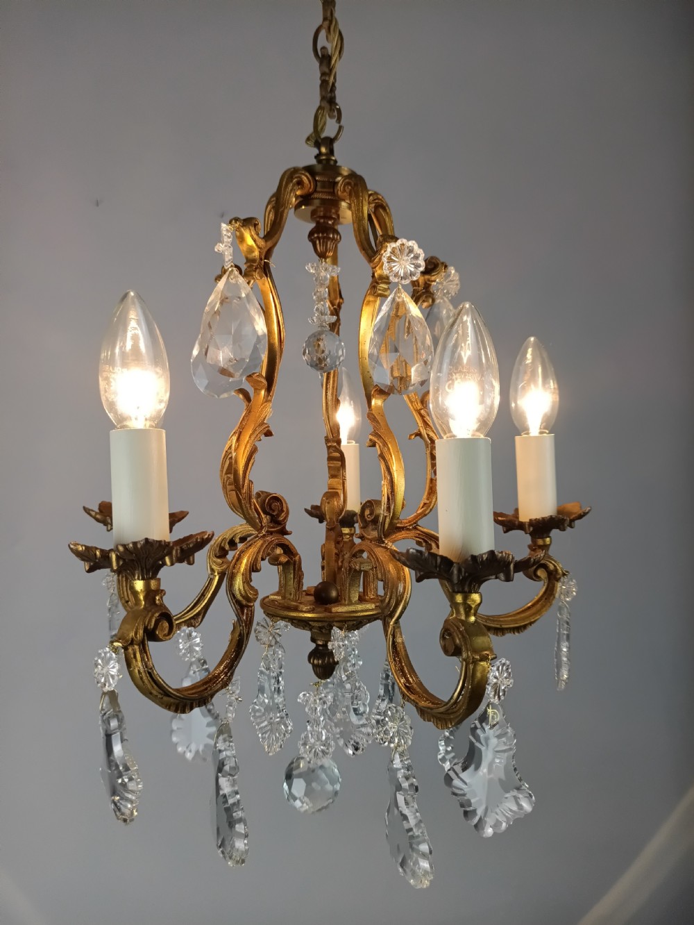 A five-arm French Rococo style chandelier featuring a gilded scrollwork frame adorned with clear faceted crystal drops and white candle sleeves. View XI