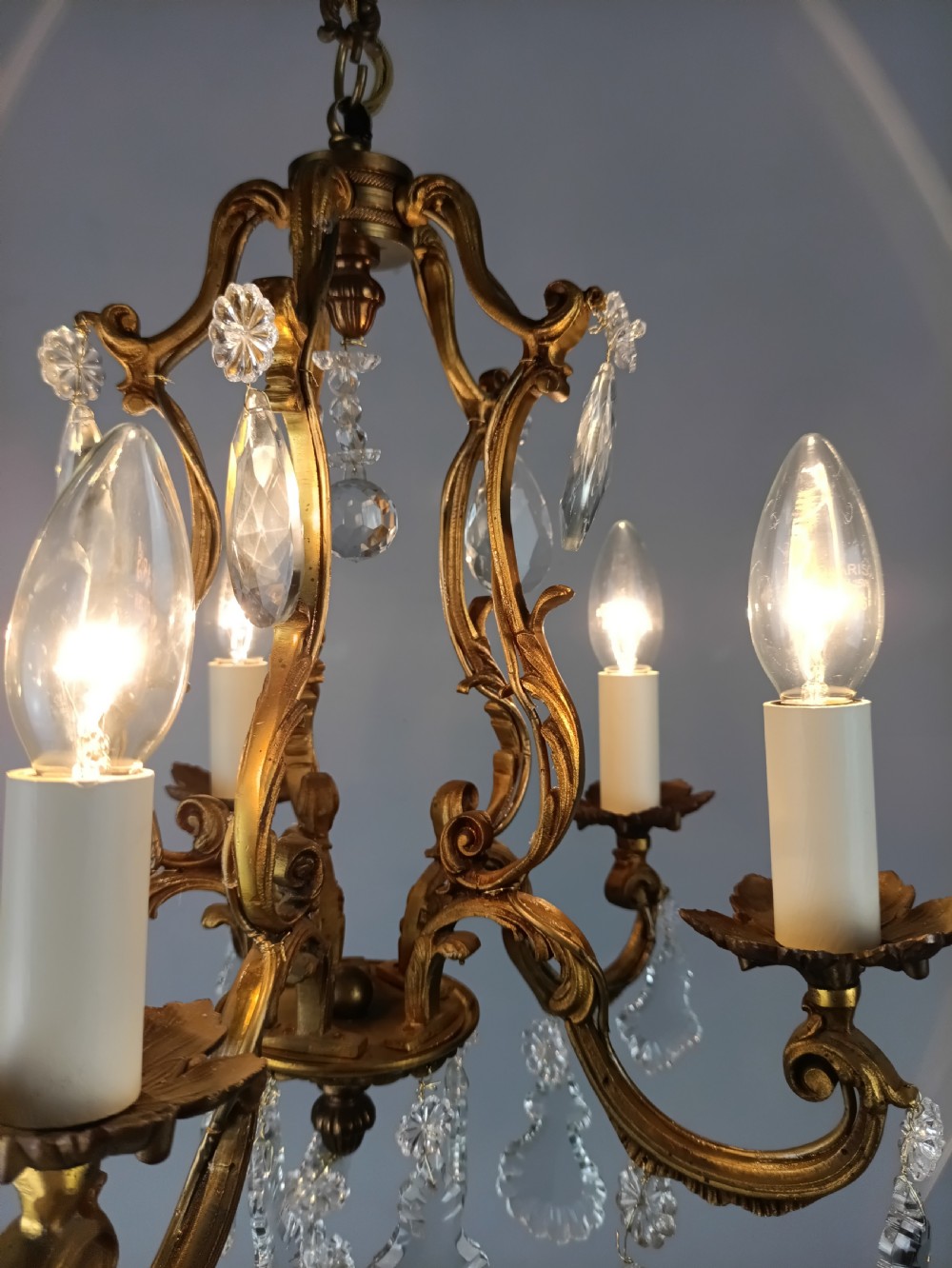 A five-arm French Rococo style chandelier featuring a gilded scrollwork frame adorned with clear faceted crystal drops and white candle sleeves. View XII