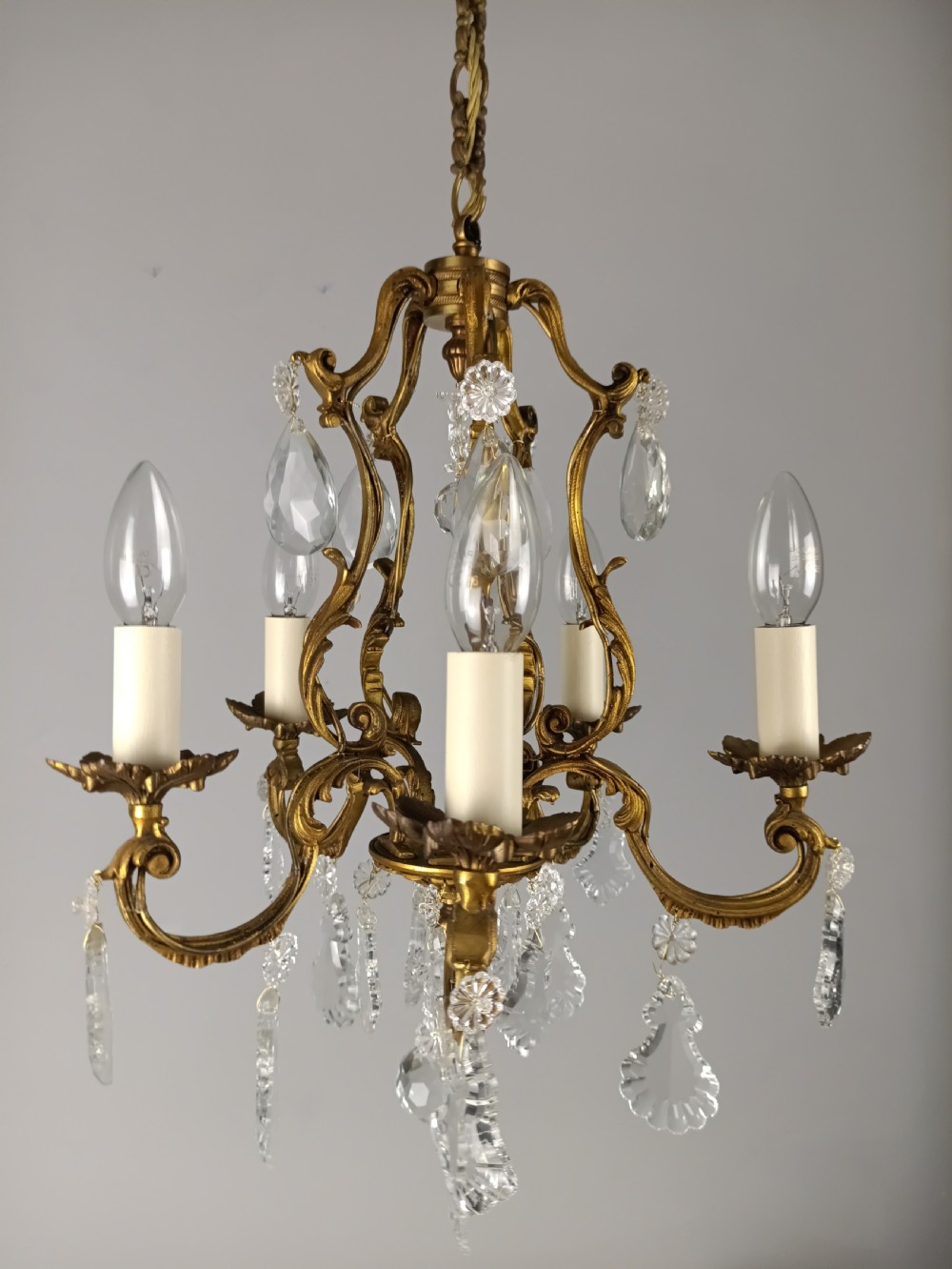 A five-arm French Rococo style chandelier featuring a gilded scrollwork frame adorned with clear faceted crystal drops and white candle sleeves. View XIII