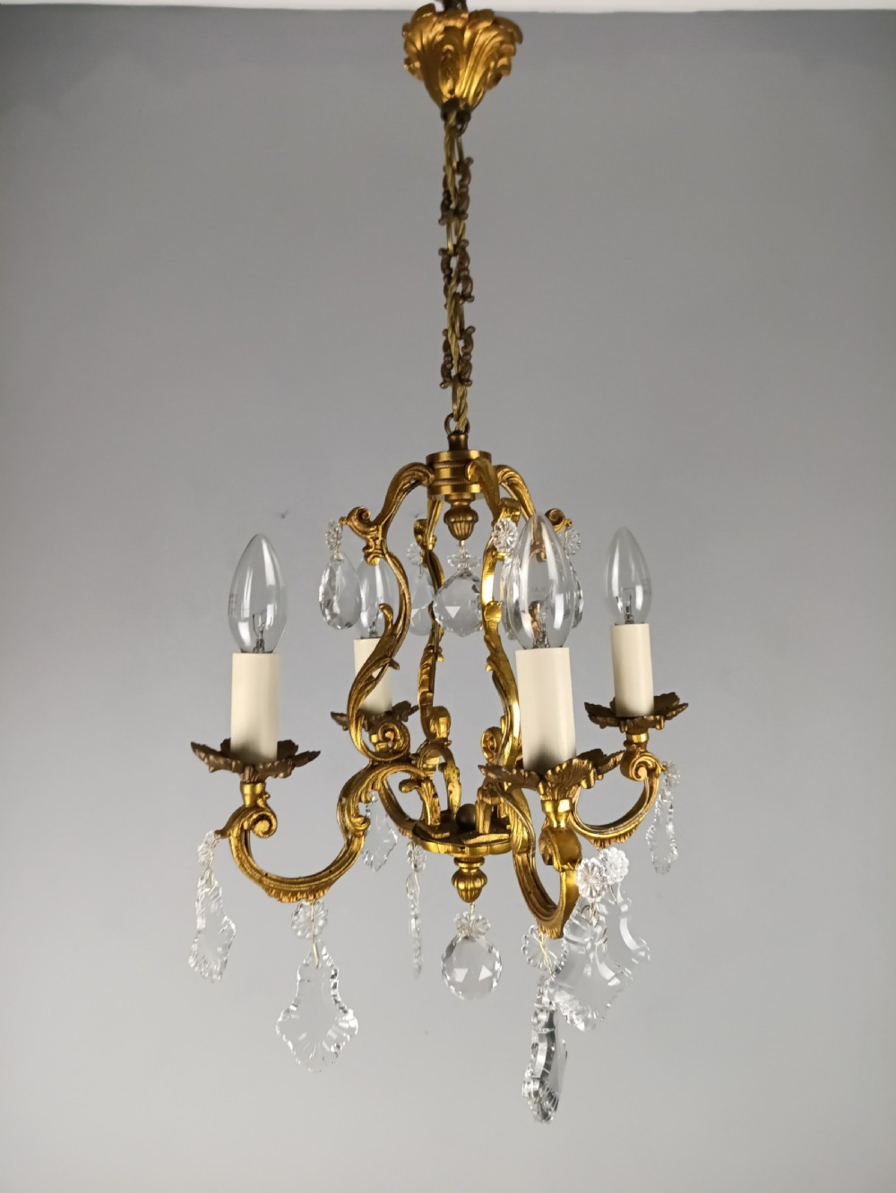 A French gilt brass four-light chandelier featuring curved scroll arms, candle sleeves, and faceted crystal droplets hanging from an ornate central frame. View II