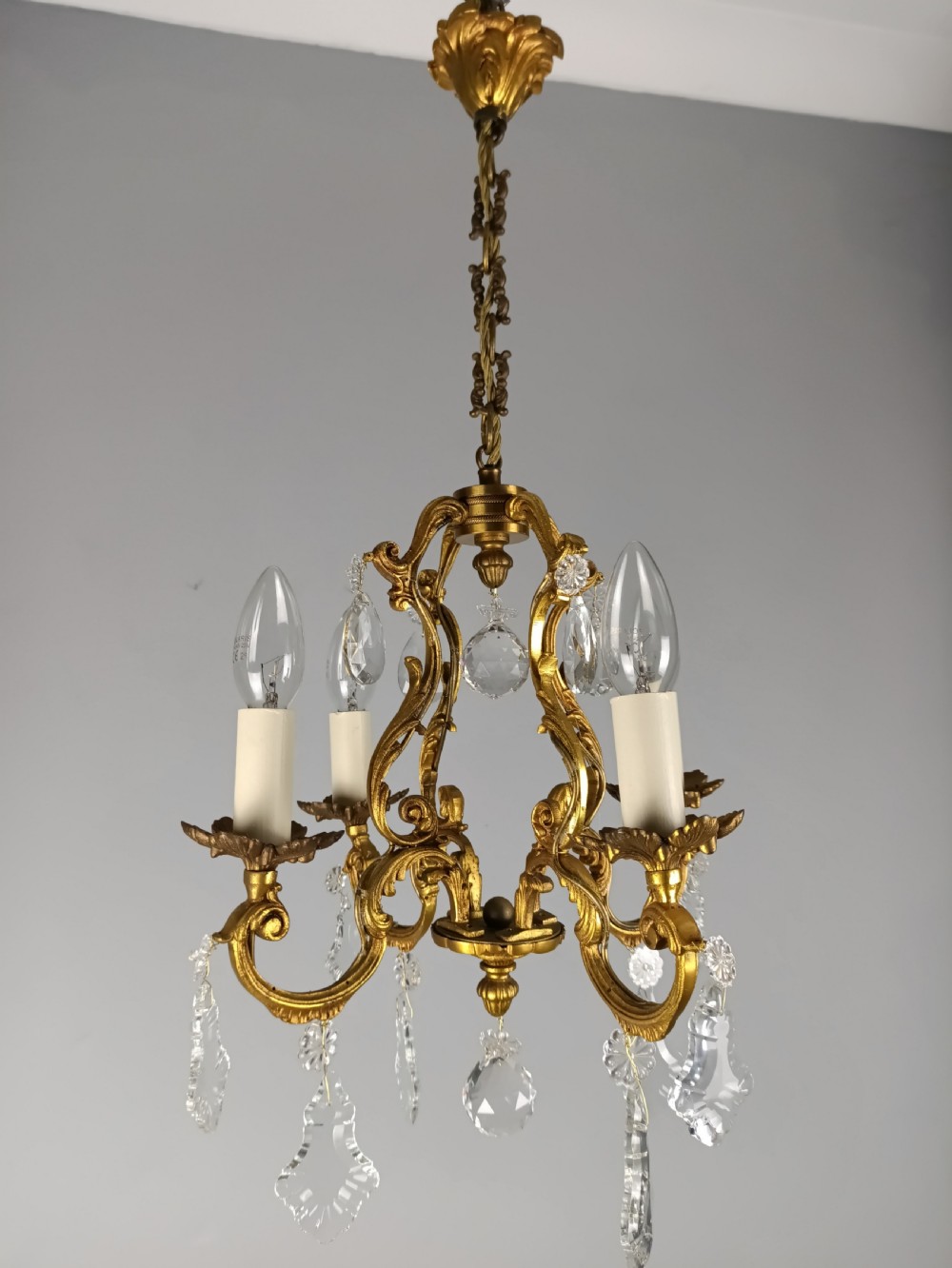A French gilt brass four-light chandelier featuring curved scroll arms, candle sleeves, and faceted crystal droplets hanging from an ornate central frame. View III