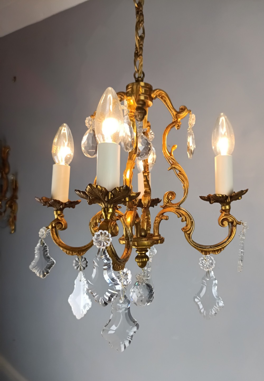 A French gilt brass four-light chandelier featuring curved scroll arms, candle sleeves, and faceted crystal droplets hanging from an ornate central frame. View IV