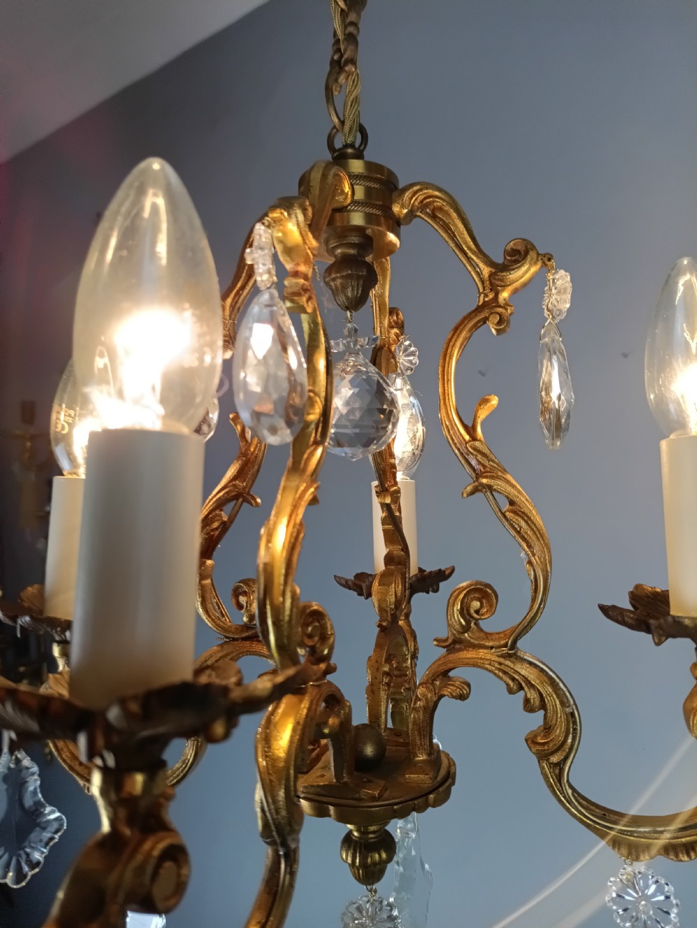 A French gilt brass four-light chandelier featuring curved scroll arms, candle sleeves, and faceted crystal droplets hanging from an ornate central frame. View VI