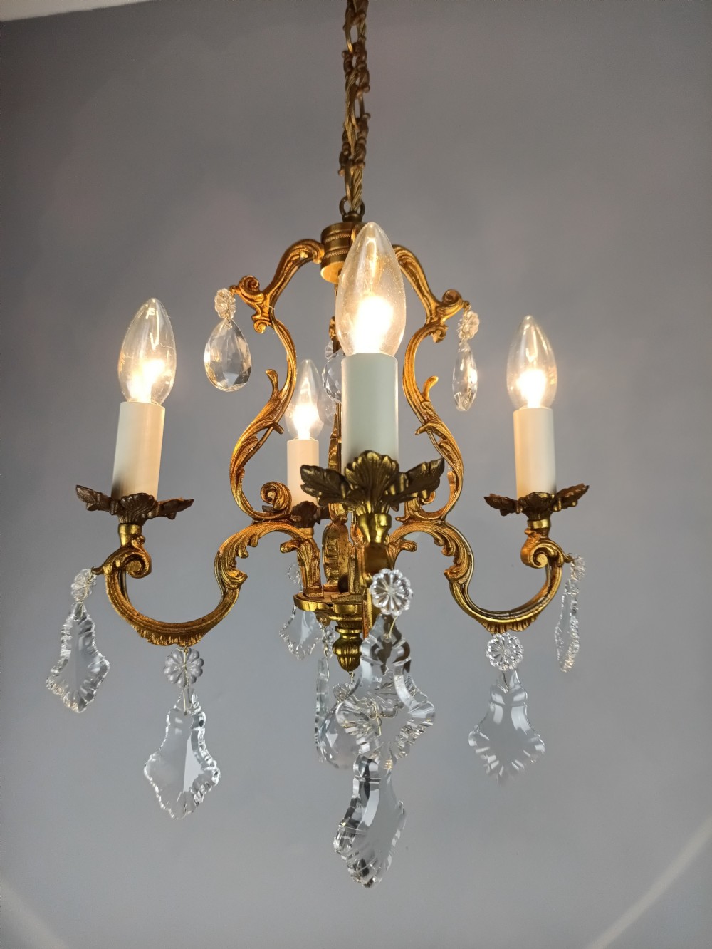 A French gilt brass four-light chandelier featuring curved scroll arms, candle sleeves, and faceted crystal droplets hanging from an ornate central frame. View VII