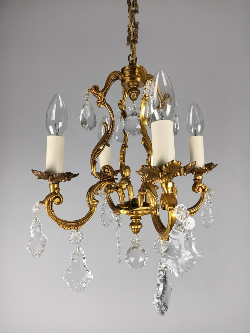 A French gilt brass four-light chandelier featuring curved scroll arms, candle sleeves, and faceted crystal droplets hanging from an ornate central frame. View X