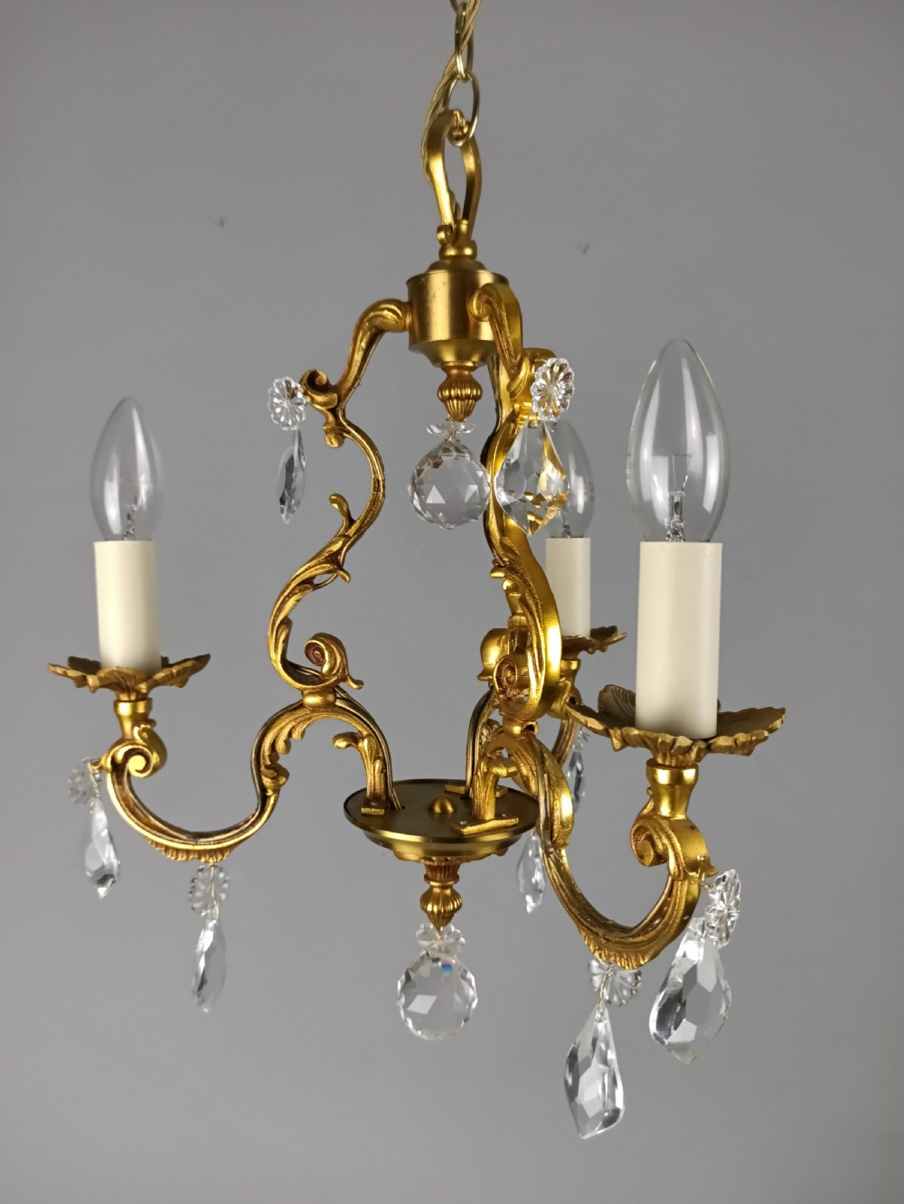 A three-arm French gilt brass chandelier decorated with scrolling foliate branches, candle sleeves, and faceted crystal glass drops.