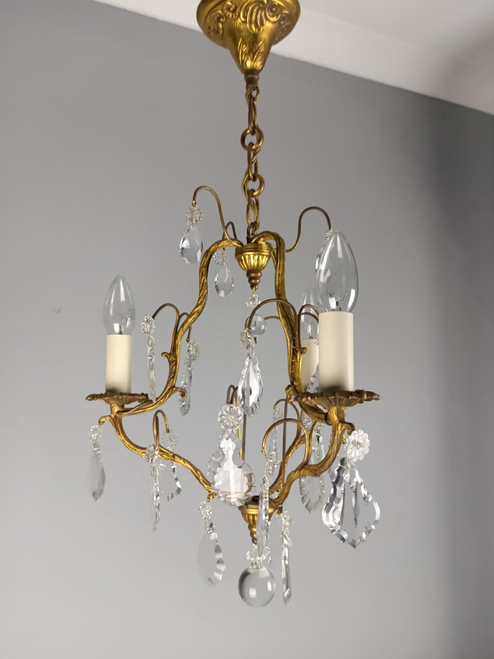 An antique French gilt brass three-light chandelier featuring curved scrolling arms, cream candle-style sleeves, and faceted crystal drops.