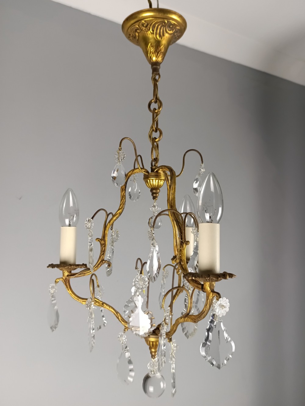 An antique French gilt brass three-light chandelier featuring curved scrolling arms, cream candle-style sleeves, and faceted crystal drops. View IV