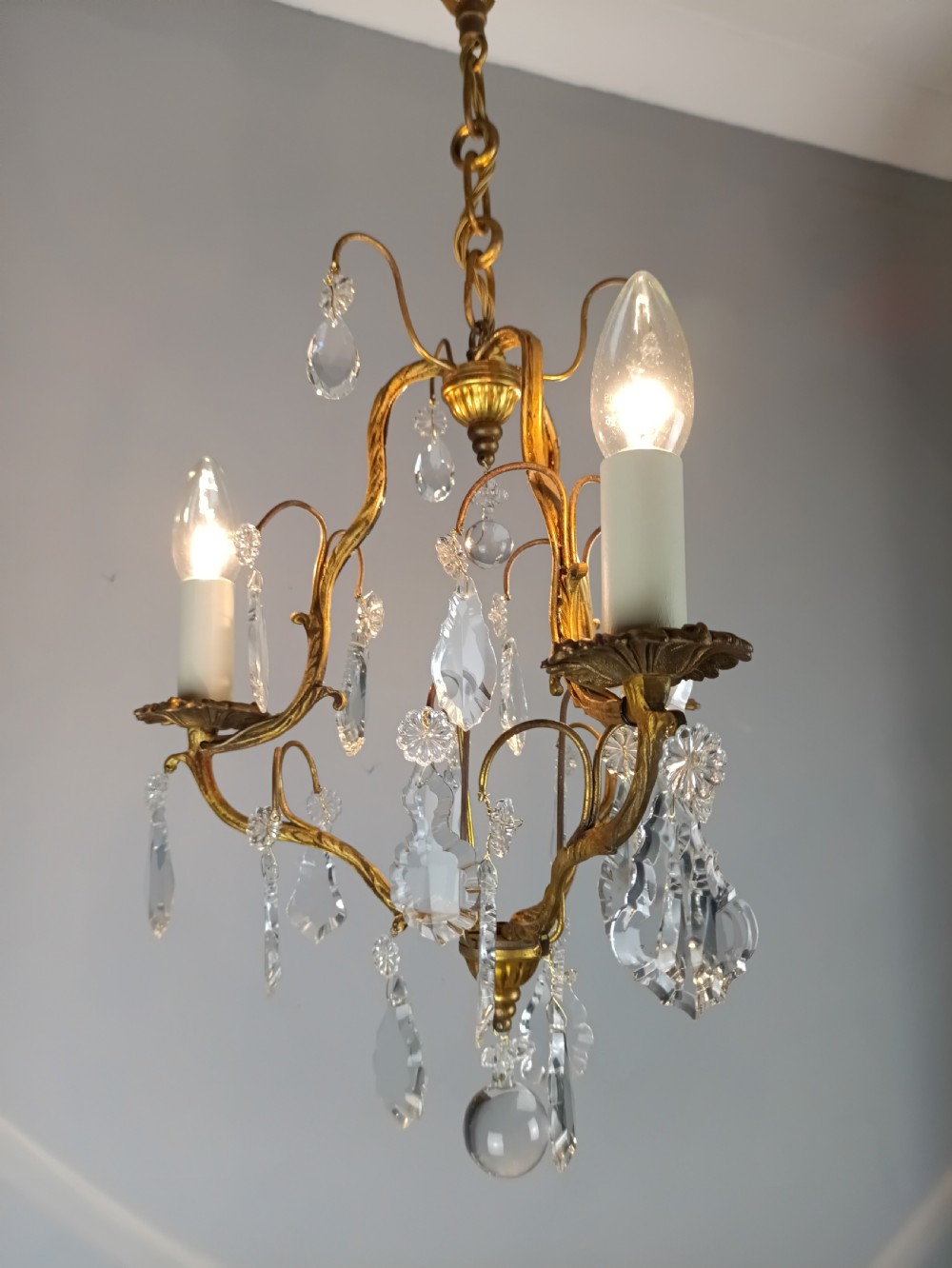 An antique French gilt brass three-light chandelier featuring curved scrolling arms, cream candle-style sleeves, and faceted crystal drops. View VI