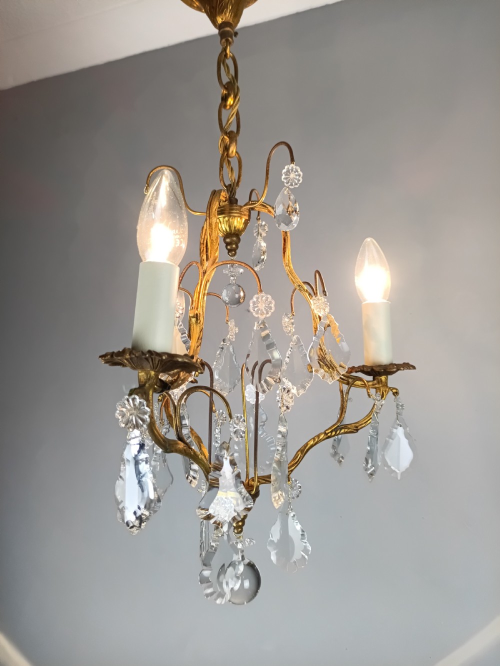 An antique French gilt brass three-light chandelier featuring curved scrolling arms, cream candle-style sleeves, and faceted crystal drops. View VII