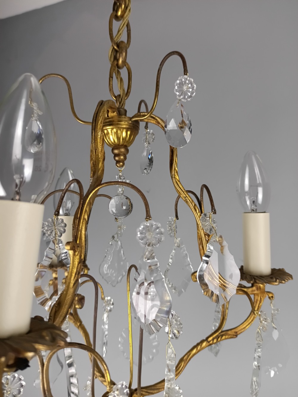 An antique French gilt brass three-light chandelier featuring curved scrolling arms, cream candle-style sleeves, and faceted crystal drops. View X