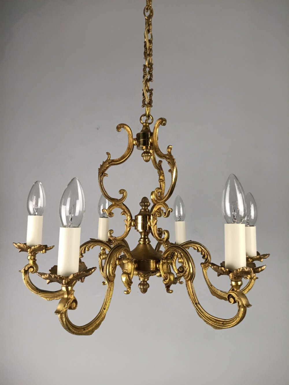 A six-arm French gilt brass chandelier featuring ornate acanthus scrollwork and white candle-style light sockets.