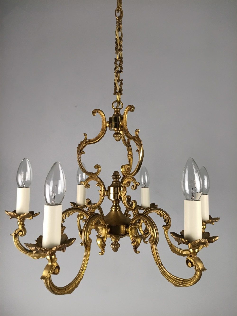 A six-arm French gilt brass chandelier featuring ornate acanthus scrollwork and white candle-style light sockets. View XII