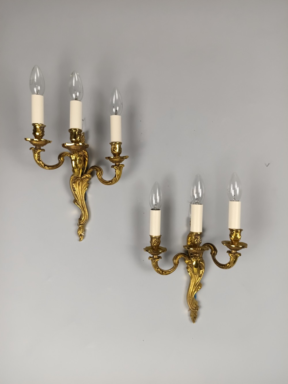 A pair of antique French Rococo wall sconces made from ornate gilded brass with three scrolling foliage-shaped arms each holding a candle-style light bulb.