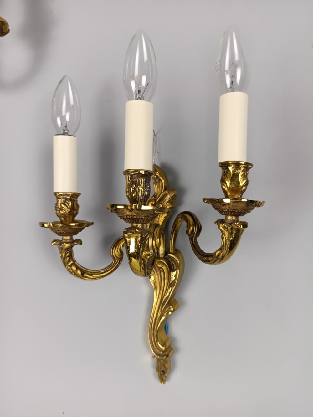 A pair of antique French Rococo wall sconces made from ornate gilded brass with three scrolling foliage-shaped arms each holding a candle-style light bulb. View II