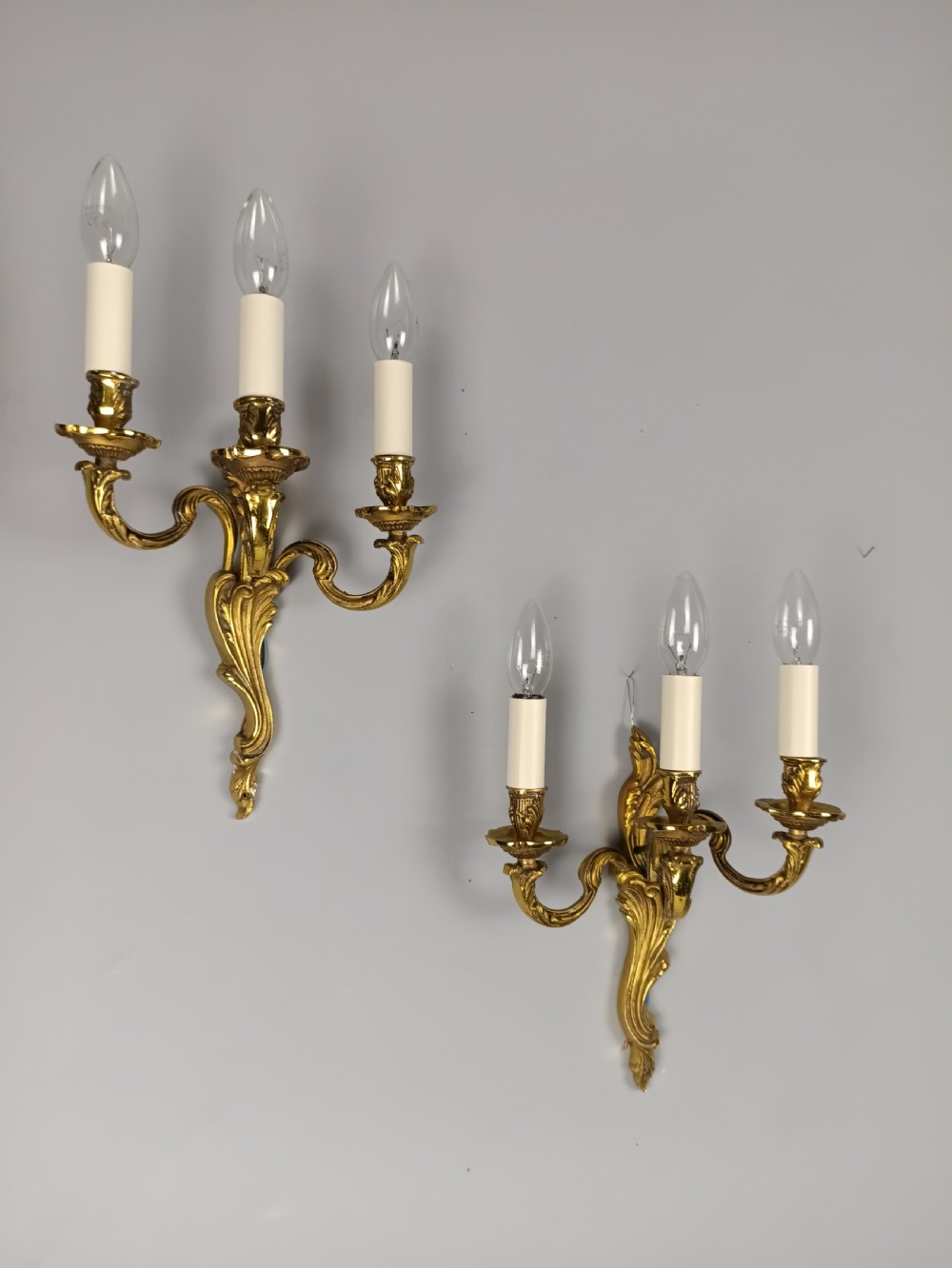 A pair of antique French Rococo wall sconces made from ornate gilded brass with three scrolling foliage-shaped arms each holding a candle-style light bulb. View IV