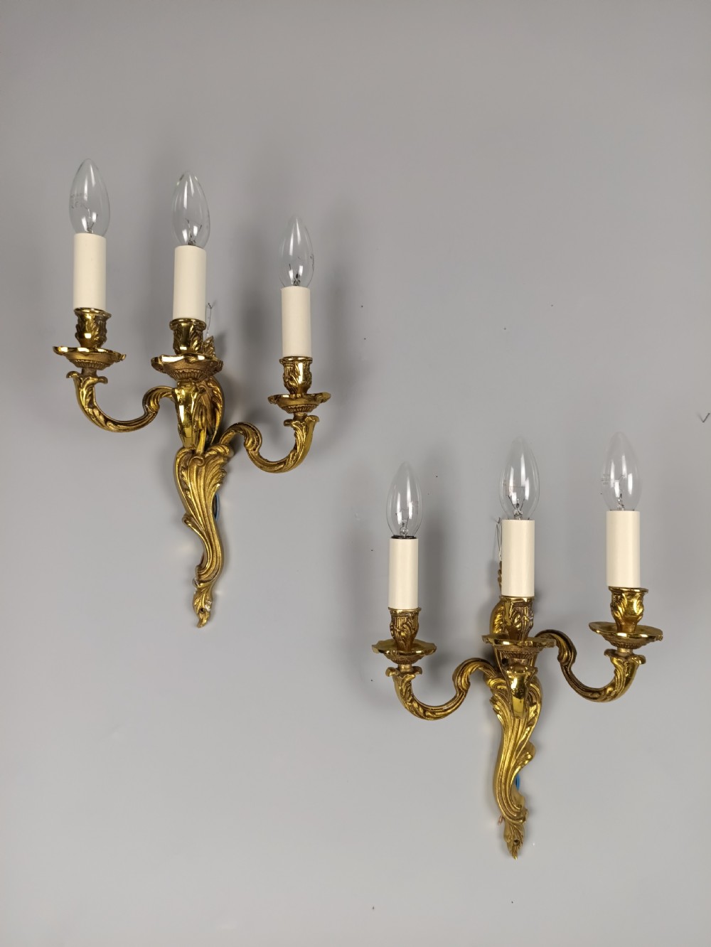 A pair of antique French Rococo wall sconces made from ornate gilded brass with three scrolling foliage-shaped arms each holding a candle-style light bulb. View VII