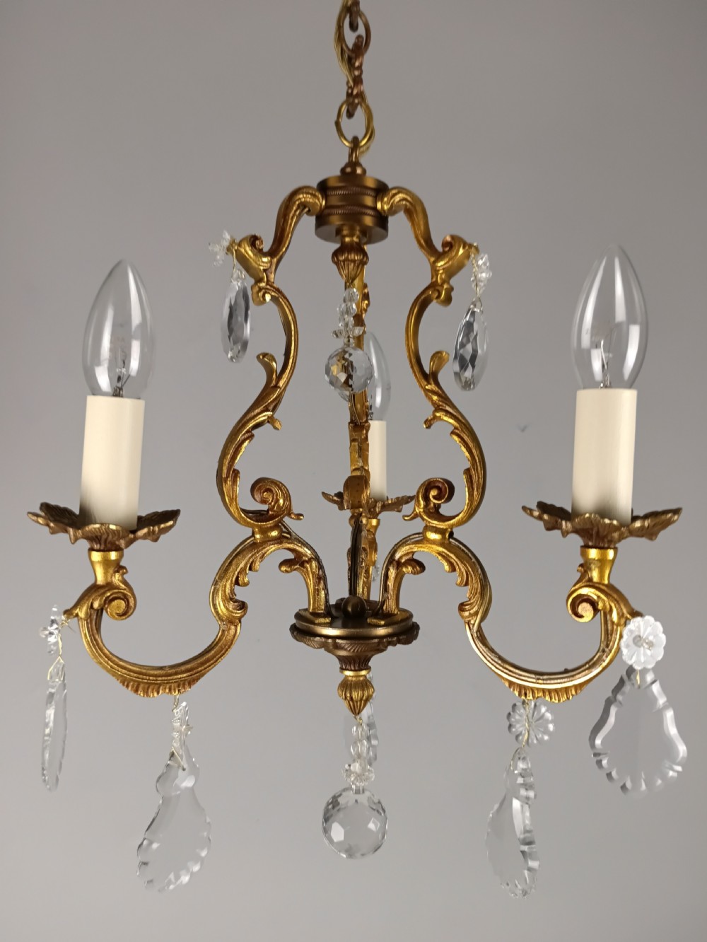 A French gilt bronze three-arm chandelier adorned with foliage scrollwork and hanging crystal droplets suspended from a ceiling chain. View IV