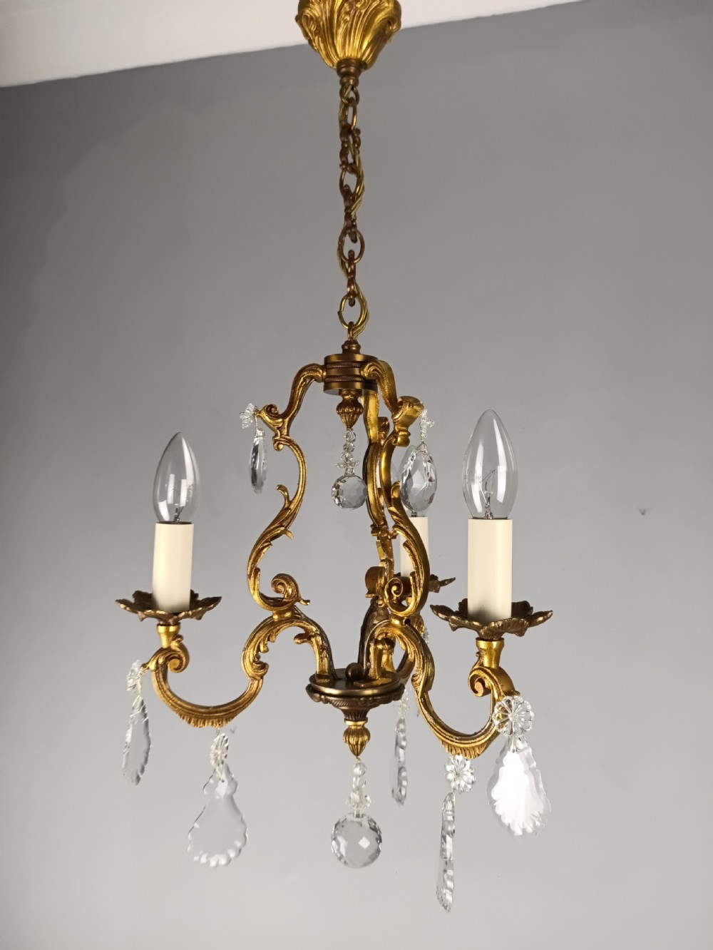 A French gilt bronze three-arm chandelier adorned with foliage scrollwork and hanging crystal droplets suspended from a ceiling chain.