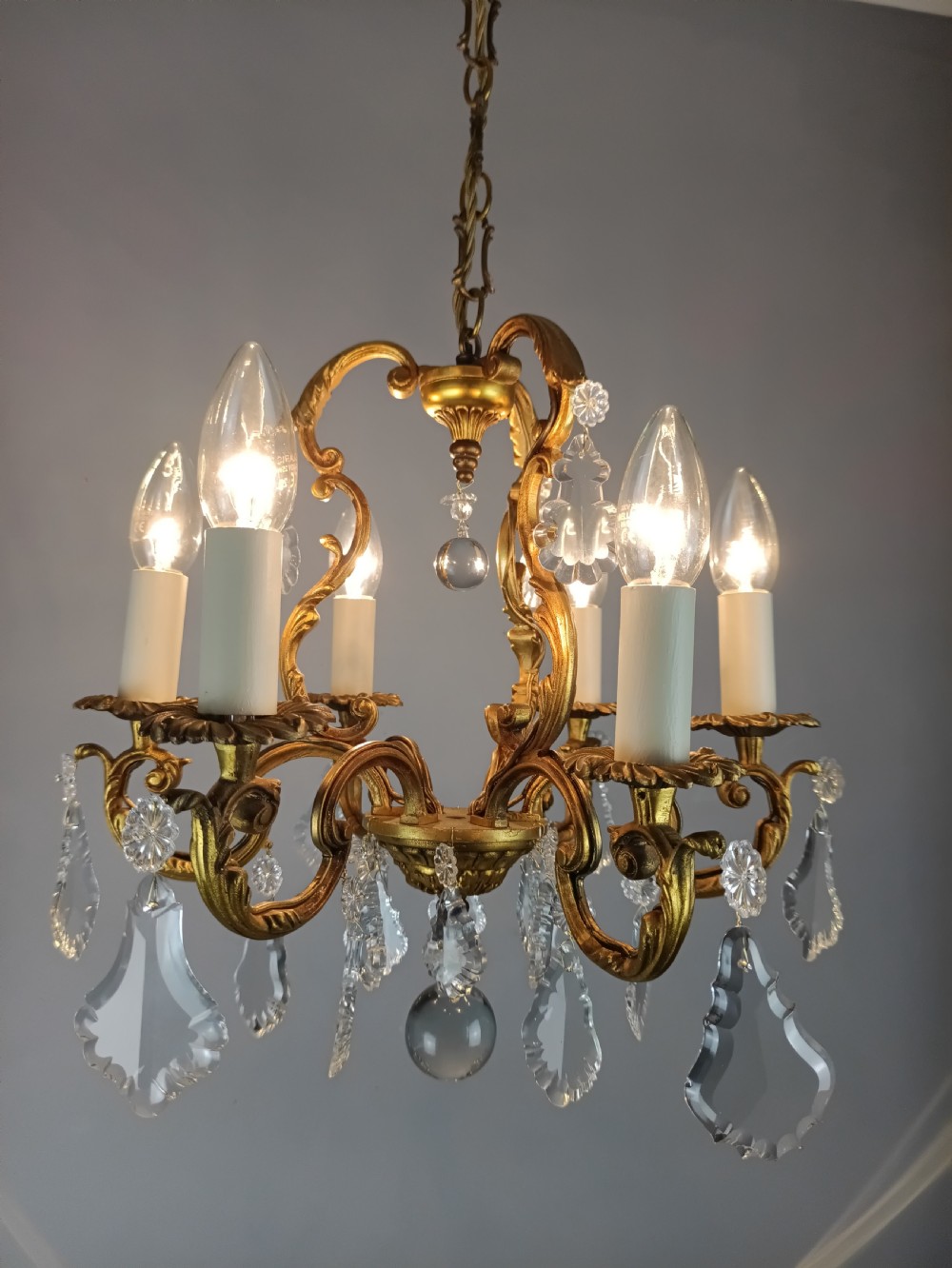 A French six-arm gilded metal chandelier featuring scrolling foliage arms, candle-style bulb holders, and hanging clear cut crystal droplets.
