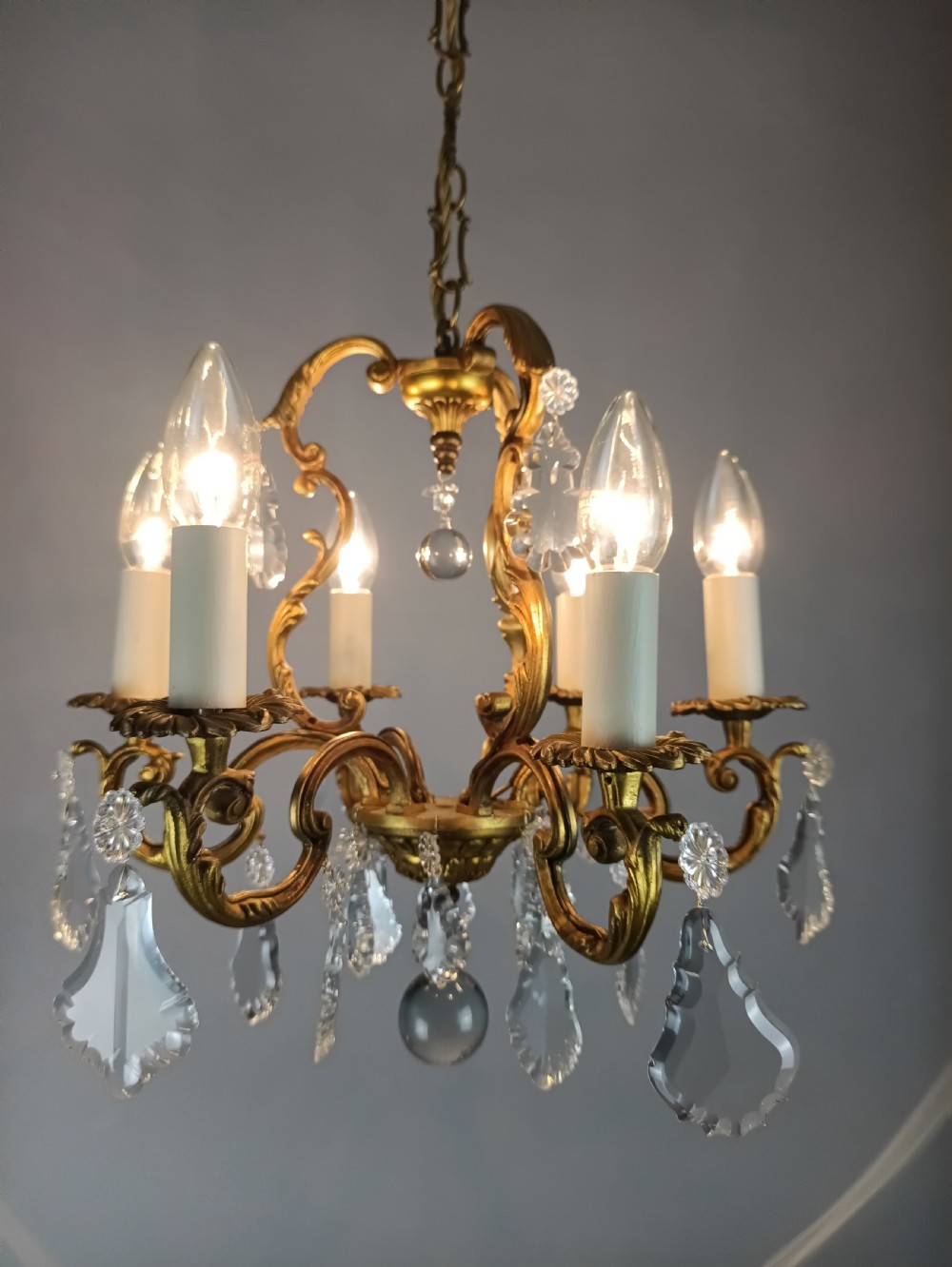 A French six-arm gilded metal chandelier featuring scrolling foliage arms, candle-style bulb holders, and hanging clear cut crystal droplets. View III