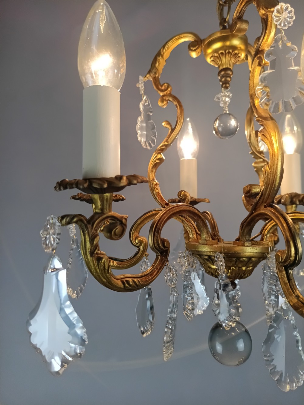 A French six-arm gilded metal chandelier featuring scrolling foliage arms, candle-style bulb holders, and hanging clear cut crystal droplets. View IV