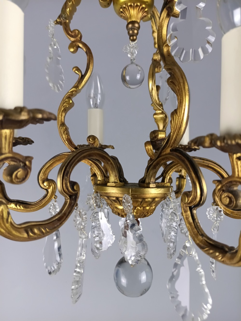 A French six-arm gilded metal chandelier featuring scrolling foliage arms, candle-style bulb holders, and hanging clear cut crystal droplets. View VI
