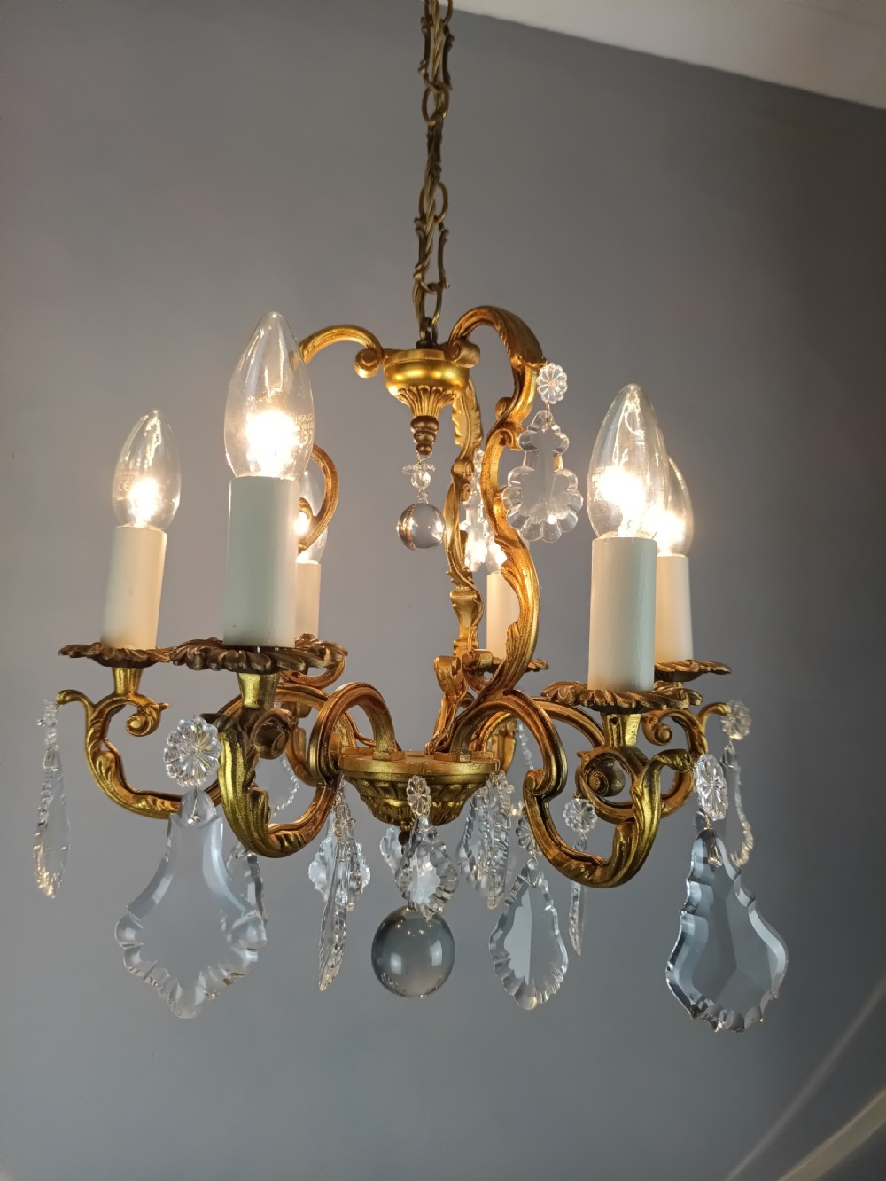 A French six-arm gilded metal chandelier featuring scrolling foliage arms, candle-style bulb holders, and hanging clear cut crystal droplets. View X