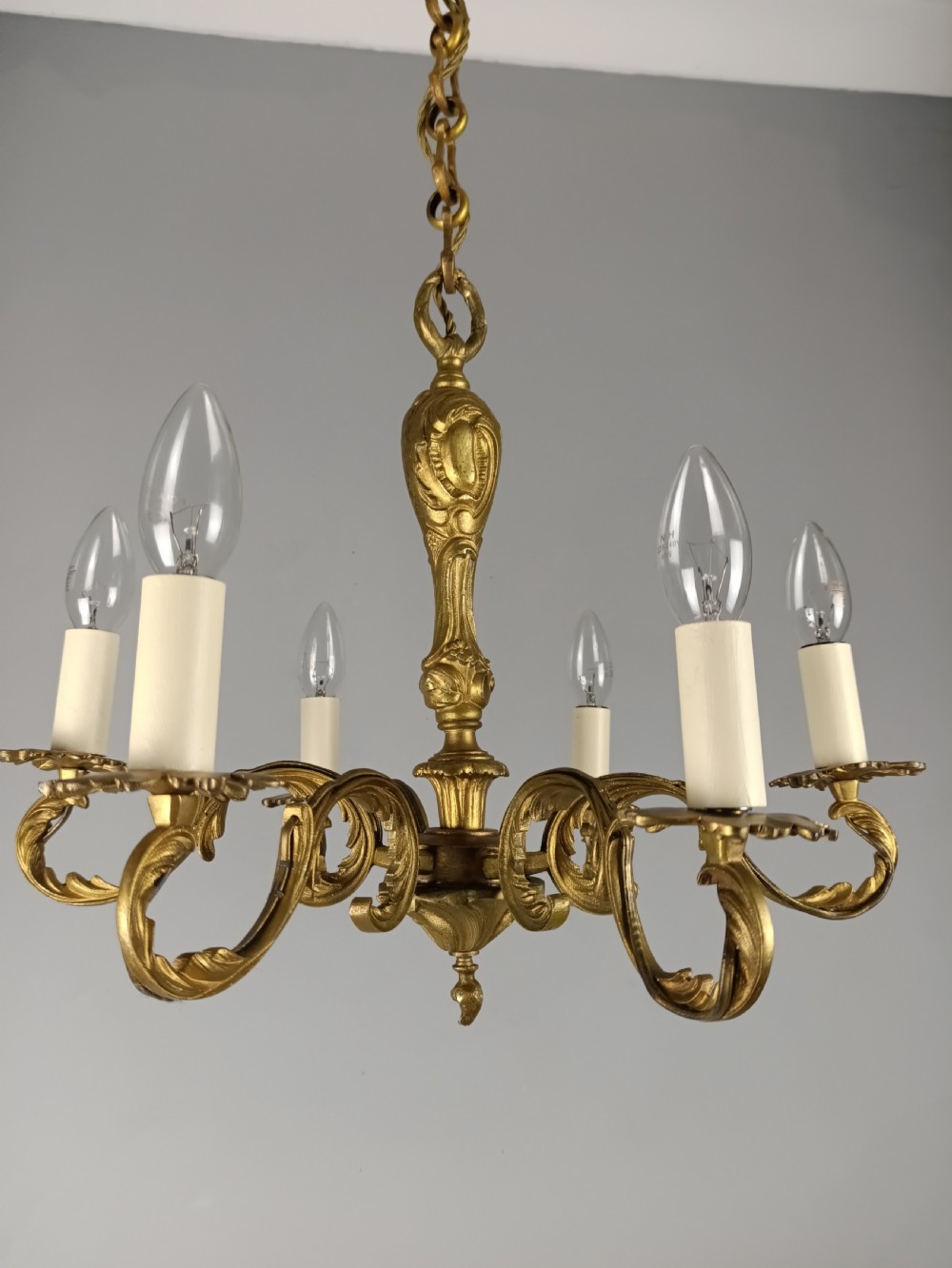 A French gilt brass six-arm chandelier featuring scrolled acanthus leaf branches, cream candle sleeves, and an ornate central column.