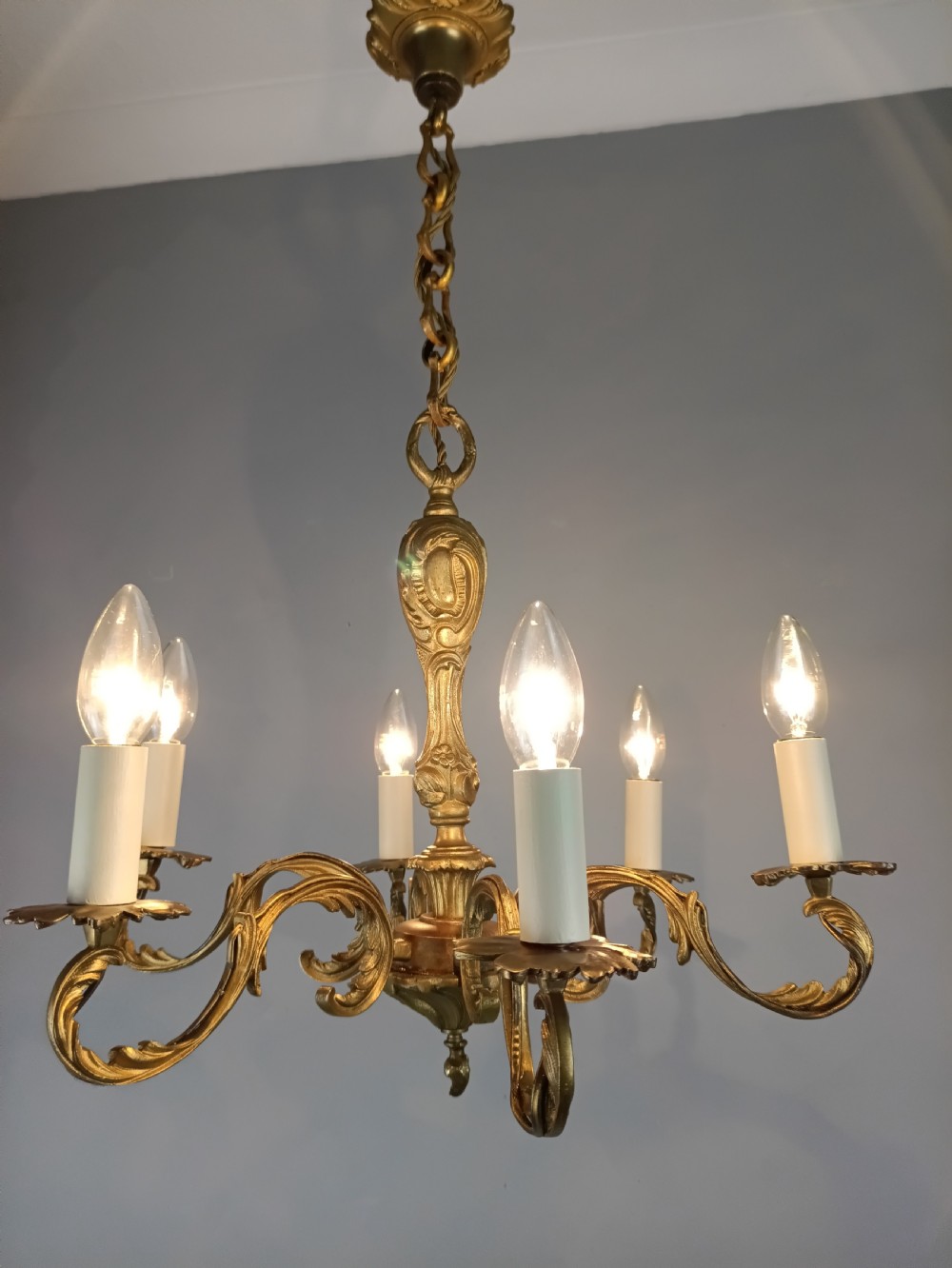 A French gilt brass six-arm chandelier featuring scrolled acanthus leaf branches, cream candle sleeves, and an ornate central column. View III