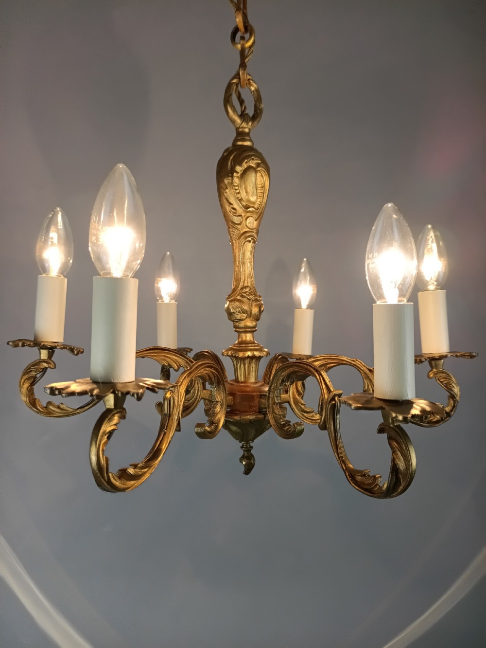 A French gilt brass six-arm chandelier featuring scrolled acanthus leaf branches, cream candle sleeves, and an ornate central column. View V